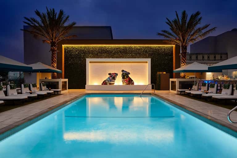 Illuminated modern resort pool at dusk with blue water, lounge areas, lit backdrop, and palm trees
