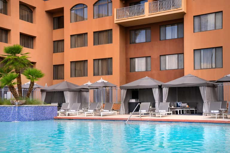 Hotel pool area with orange building facade, gray cabana umbrellas, and palm landscaping