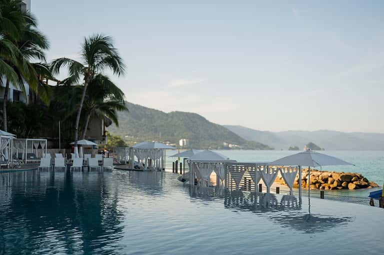 Infinity pool merging with ocean, palm trees, mountain views, and white cabanas at sunset
