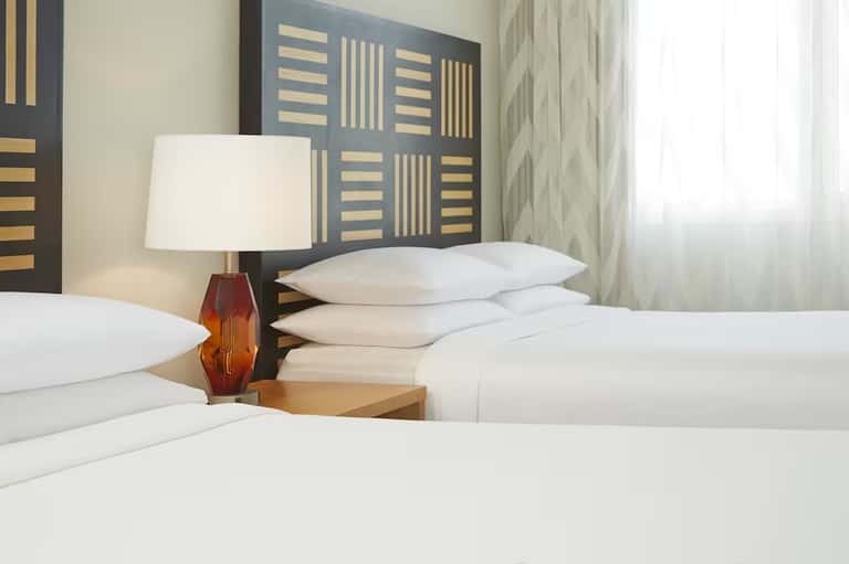 Hotel room detail showing white pillows, decorative headboard, bedside lamp, and curtains