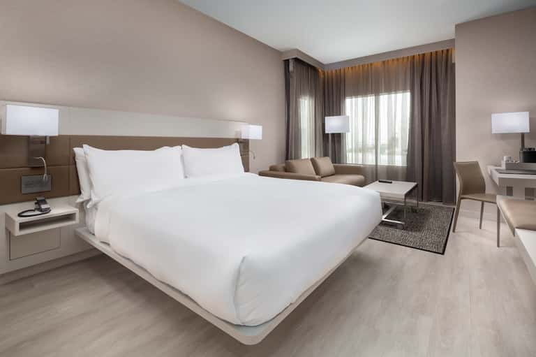 Modern hotel bedroom with king bed, seating area, and floor-to-ceiling windows