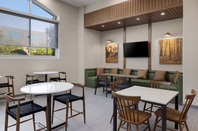Modern hotel lounge with round tables, wooden chairs, green seating area, TV, and large windows