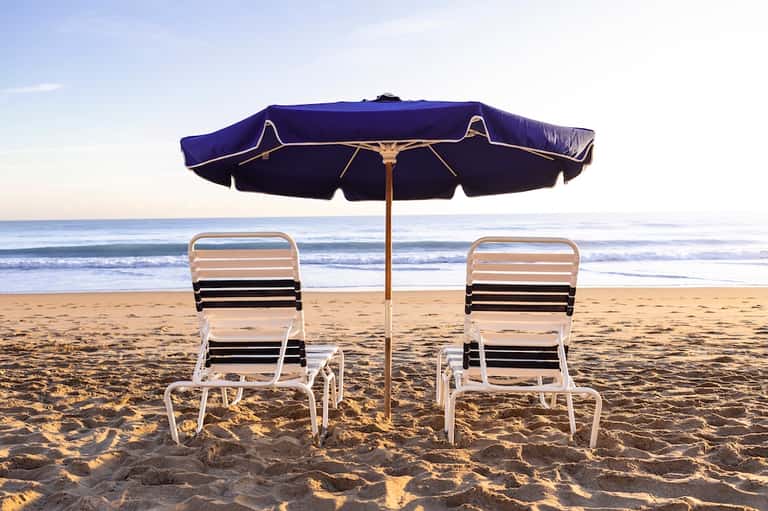 Two lounge chairs under navy beach umbrella on sandy beach with calm ocean waves in background