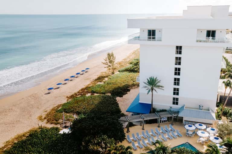 Beachfront resort building overlooking sandy beach, ocean, blue lounge chairs, white umbrellas, and palm landscaping