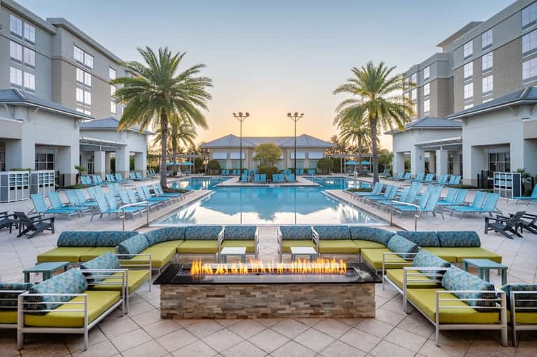 Resort pool deck at dusk with turquoise loungers, lime cushioned seating, fire pit, palm trees, and modern buildings