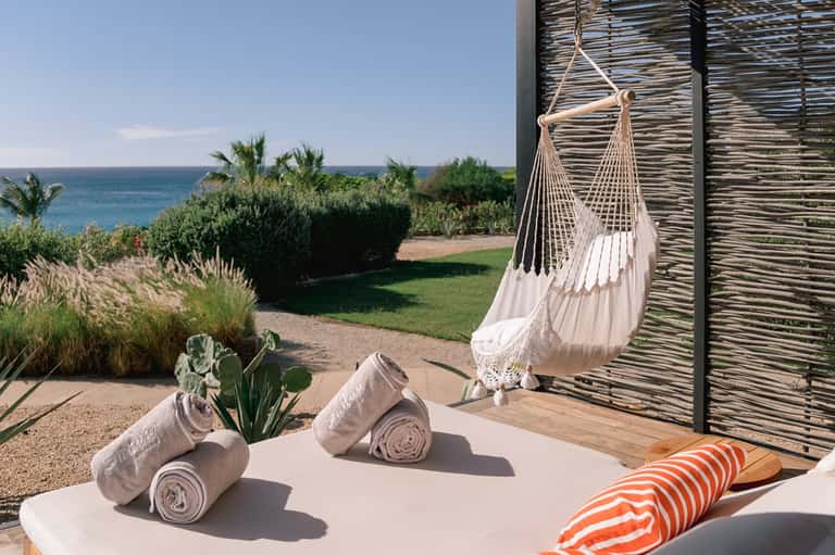 Beachfront terrace with hanging white hammock, rolled towels, ocean view, palm trees, and privacy screens