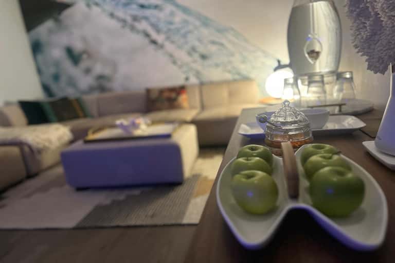 Hotel suite detail showing fresh green apples in bowl, glass container, and blurred seating area in background