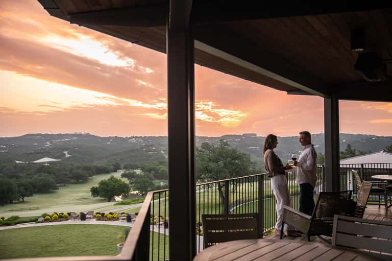 Couple on sunset balcony overlooking green valley, lake, and rolling hills at dusk