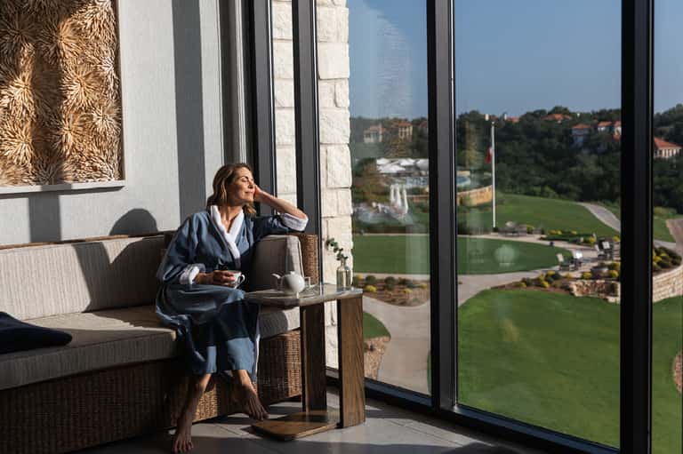 Woman in robe on seating area with expansive windows overlooking green hillside landscape and distant valley views