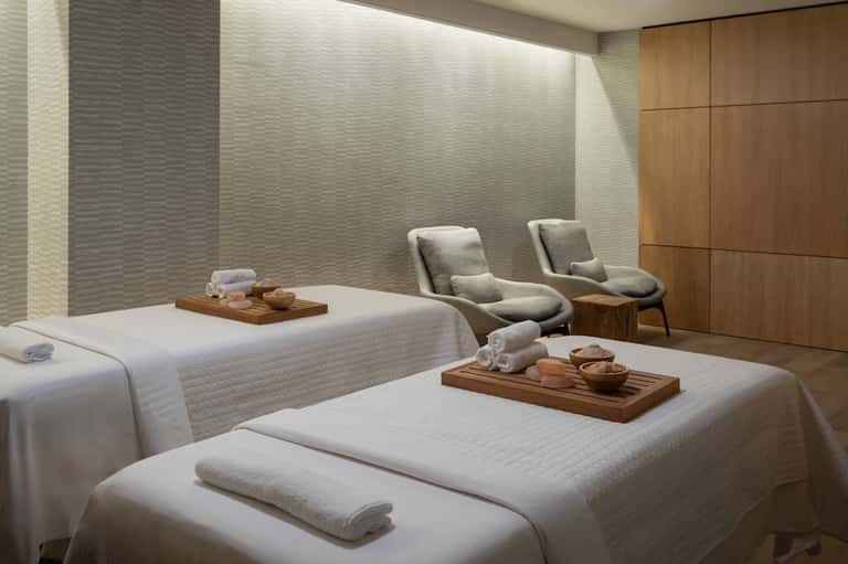 Spa treatment room with massage beds, neutral tones, wooden cabinetry, and relaxation seating