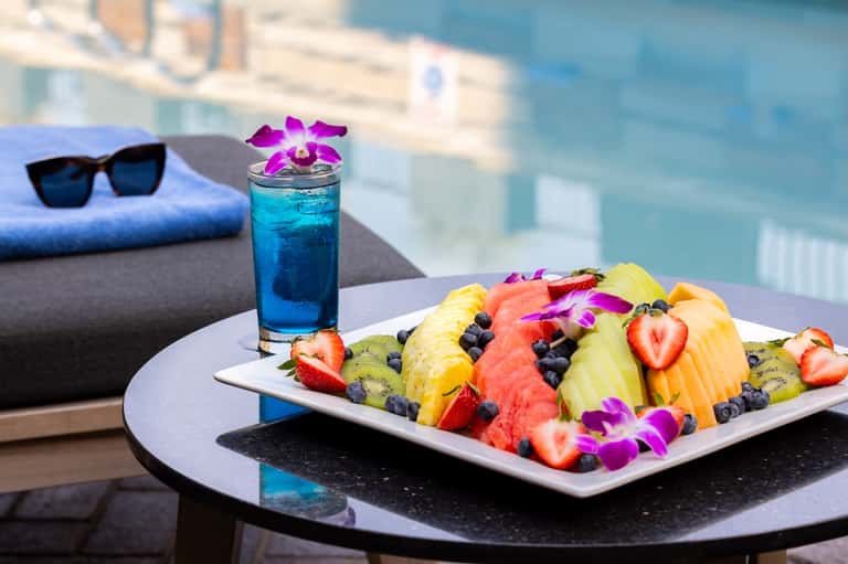 Poolside table with colorful fruit plate, blue cocktail, pink orchid, and city skyline backdrop