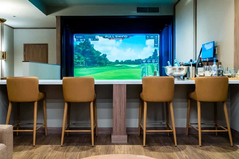 Golf simulator bar with four tan leather stools facing large golf game screen display