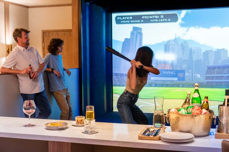 Interactive entertainment room with virtual baseball game, people playing, beverages, and snacks on counter