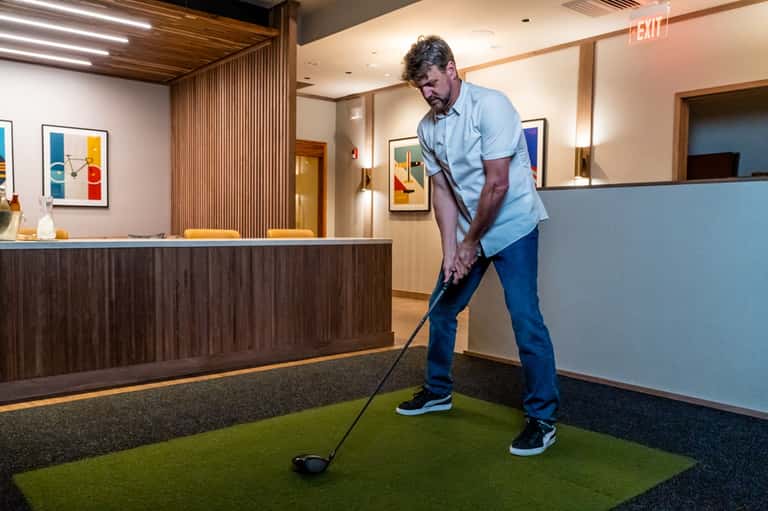 Man practicing golf swing on indoor putting green near hotel lobby with reception desk