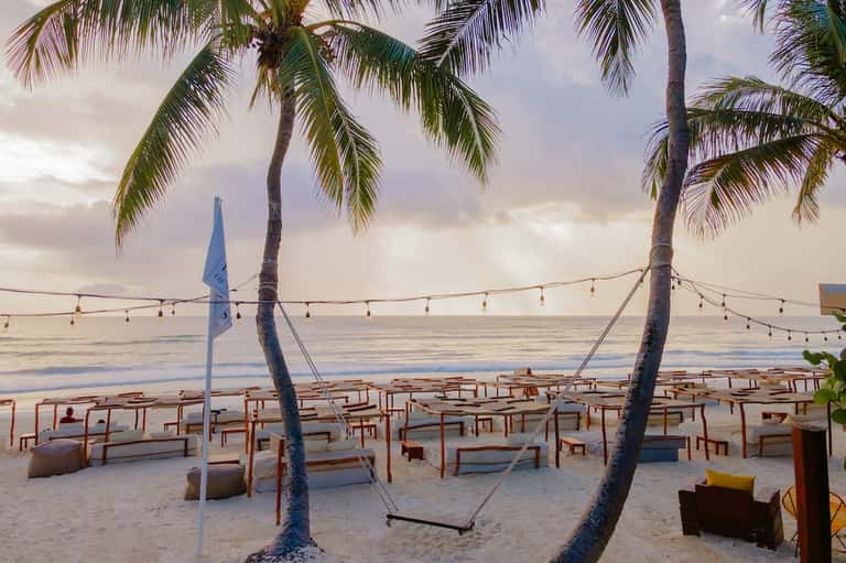 Beach lounge setup at dusk with wooden chairs, palm trees, string lights, and calm ocean horizon view