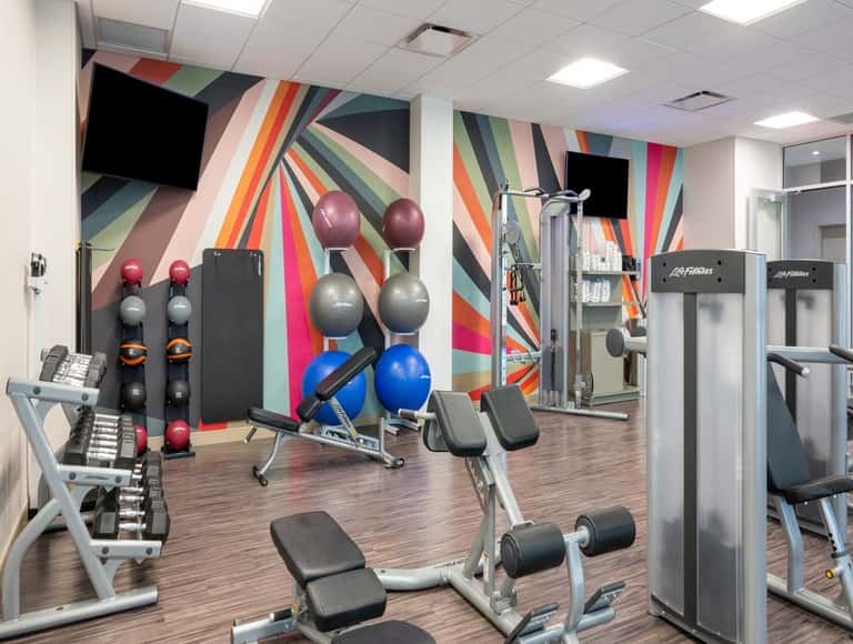 Fitness center with colorful geometric mural, dumbbells, exercise equipment, and TVs