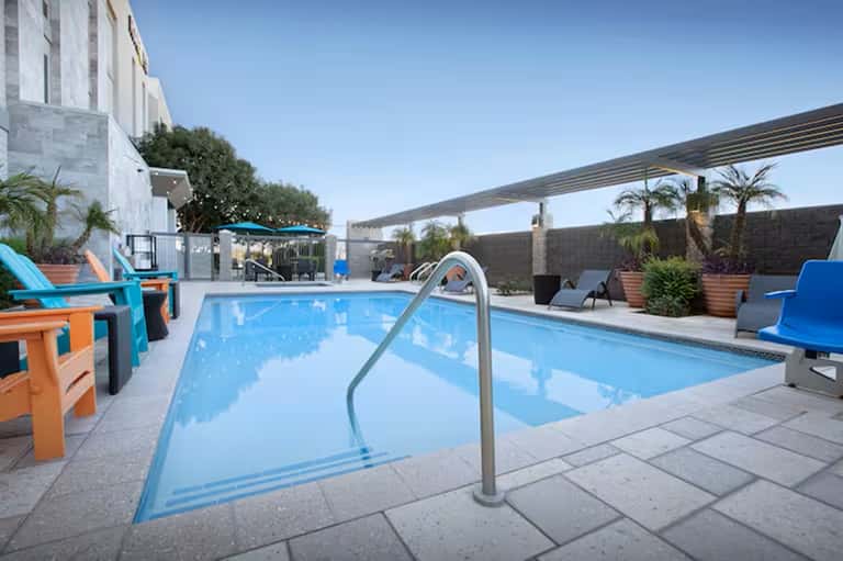 Contemporary resort pool with blue water, colorful lounge chairs, shaded pergola, and modern building architecture.
