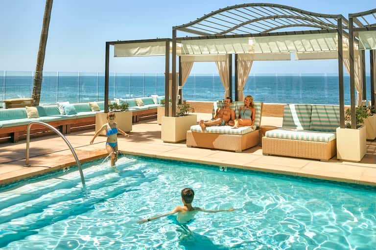 Beachfront resort pool with pergola shade structures, lounge seating, ocean view, and family enjoying water