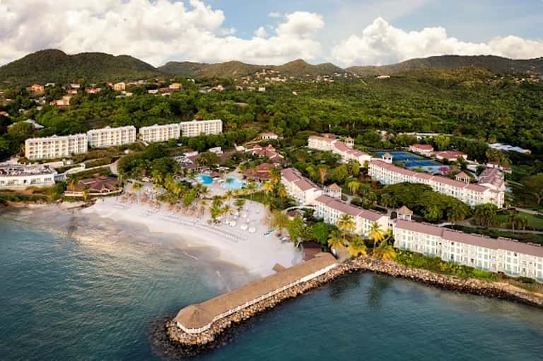 Beachfront resort aerial view with white sand, pools, palm trees, and mountainous backdrop