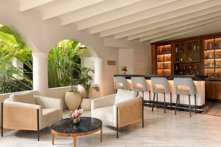 Elegant lobby lounge with wicker chairs, arched columns, tropical plants, and illuminated bar in background