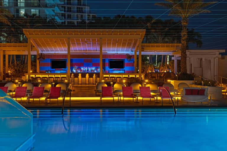 Nighttime resort pool deck with blue-lit water, illuminated lounge pavilion, red chairs, and entertainment venue backdrop