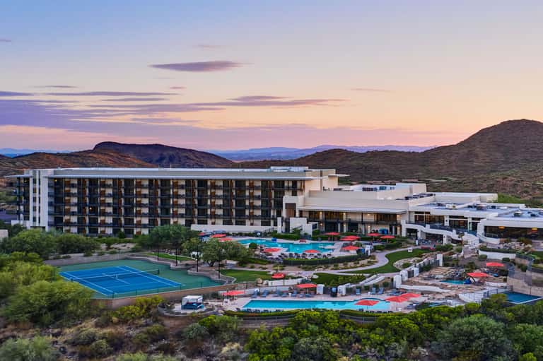 Desert resort at dusk with modern architecture, multiple pools, tennis court, and mountain views