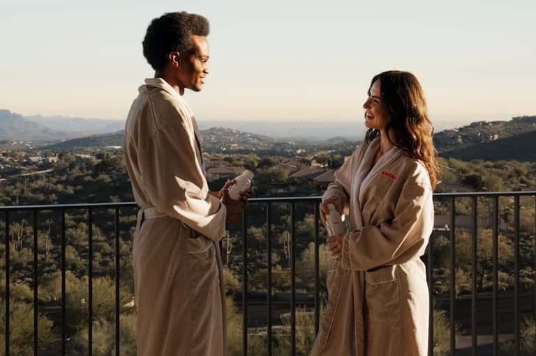 Two people in robes on a hillside terrace overlooking a scenic valley landscape