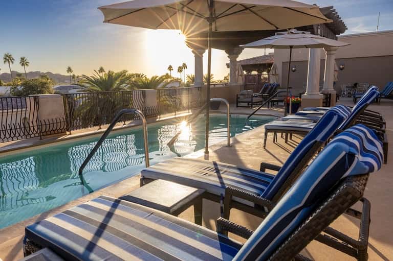 Rooftop pool deck with blue lounge chairs, umbrellas, mountain views, and desert landscape at sunset