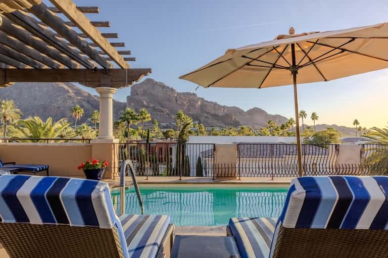 Resort pool with mountain views, umbrellas, striped lounge chairs, and pergola shade structure