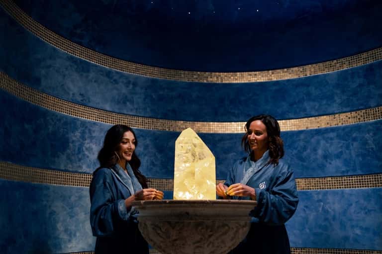 Two spa attendants in blue robes standing by illuminated crystal in luxurious tiled spa room