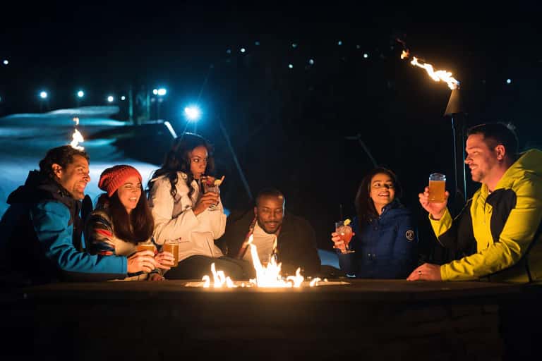 Group of friends sitting around a fire pit by the pool at night celebrating with drinks
