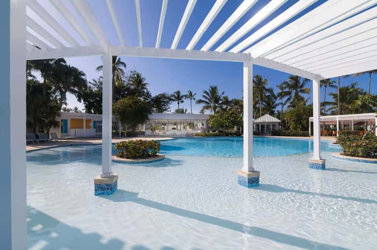 Luxury resort pool with white pergola, shallow wading area, palm landscaping, and modern white cabanas