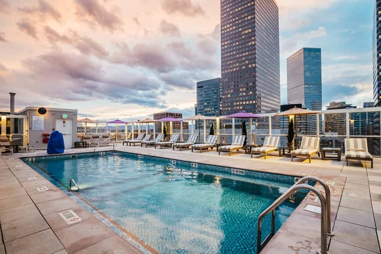 Rooftop pool deck at sunset with city skyline, lounge chairs, umbrellas, and urban backdrop