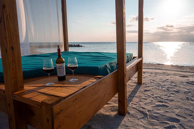 Beachfront cabana with turquoise cushions, wine glasses, and ocean sunset view with sheer curtains