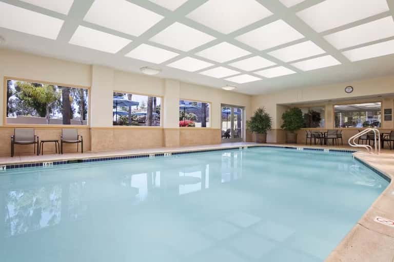 Indoor heated swimming pool with skylights, large windows, lounge chairs, and tropical plants