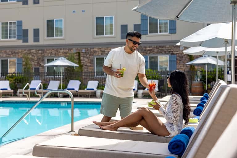 Couple enjoying beverages poolside under umbrellas at resort with building and landscaping backdrop