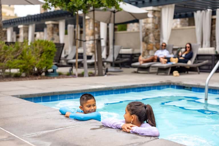 Resort pool with children in water, lounge seating area, cabanas, and parents watching