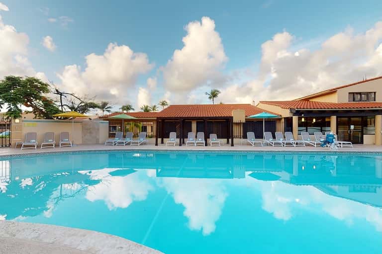 Resort pool reflecting sky with cabanas, lounge chairs, palm trees, and Spanish-style buildings