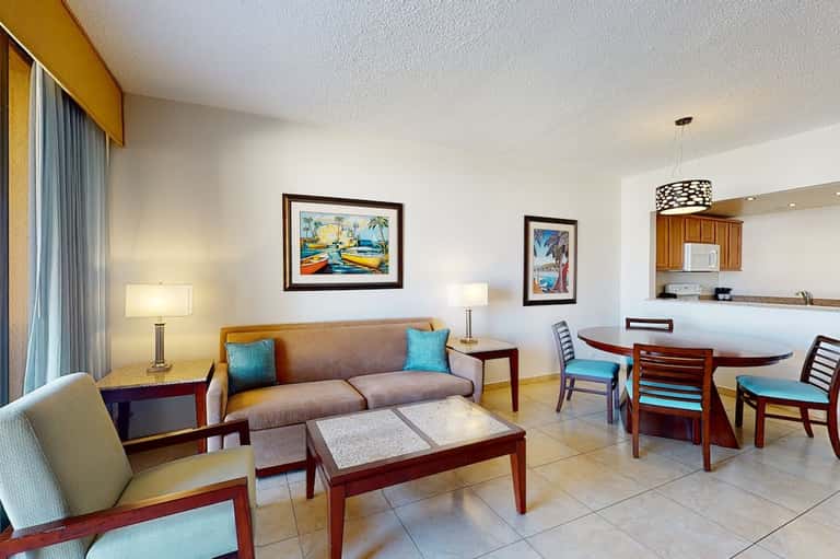 Hotel suite living area with sofa, dining table, kitchenette, and tropical-themed artwork