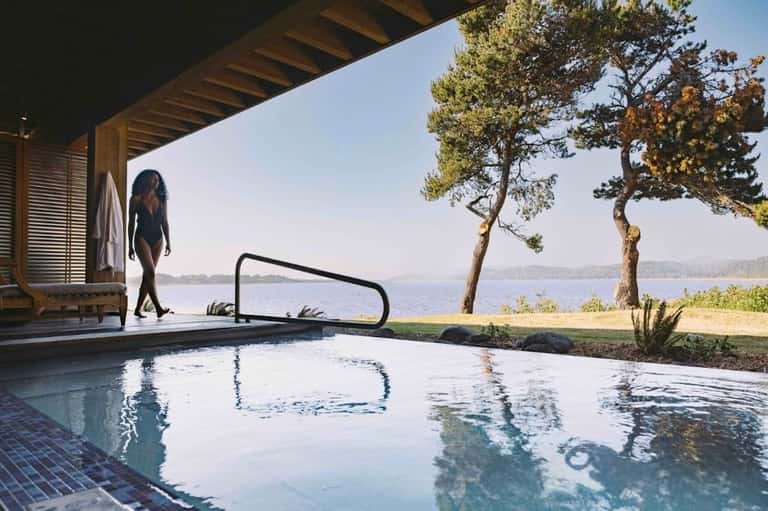 Woman in swimwear approaching natural hot spring pool overlooking lake and forested landscape
