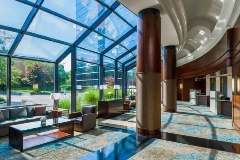 Contemporary hotel atrium with glass ceiling, wooden columns, floor seating, and outdoor garden views