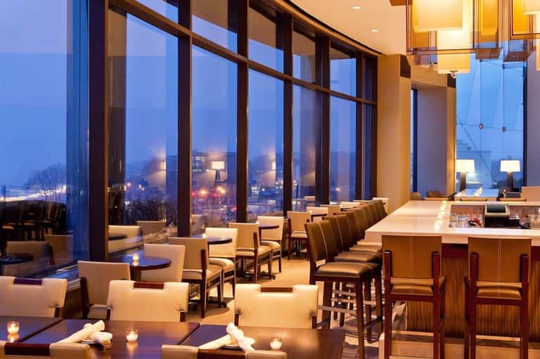 Upscale dining lounge with floor-to-ceiling windows, city night views, contemporary furniture, and bar