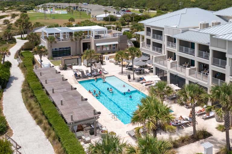 Aerial view of resort with outdoor pool, lounge chairs, palm trees, and multi-unit buildings