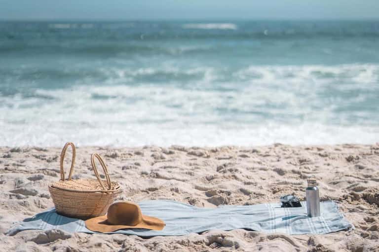 Beach picnic setup with wicker basket, straw hat, and blanket on sand overlooking ocean waves