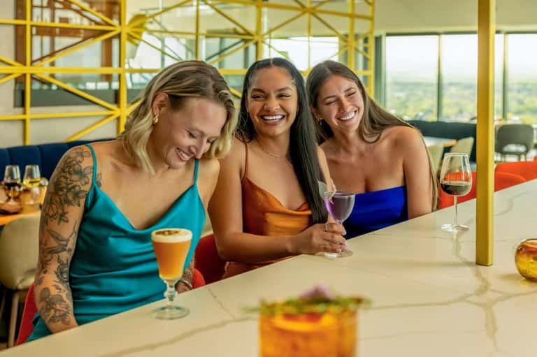 Three women at indoor bar with yellow metal railings, cocktails, and panoramic landscape views