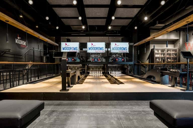 Modern bowling alley with three lanes, wood ceiling beams, neon signage, and casual seating
