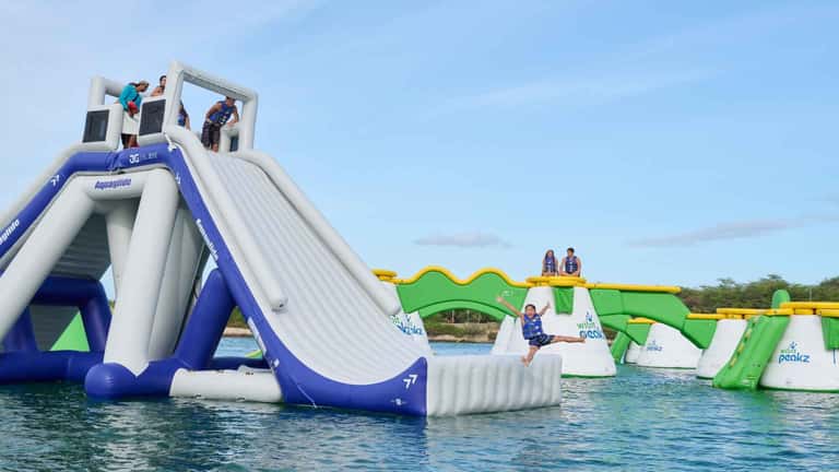 Inflatable water park with blue and green slides, obstacles, and guests enjoying activities in shallow ocean water