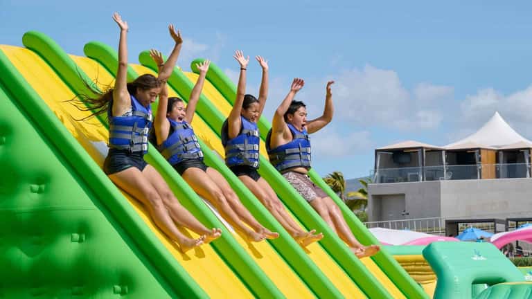 Four people sliding down giant inflatable water slide wearing life jackets with arms raised in joy