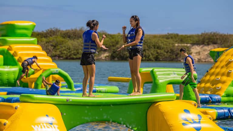 Children playing on inflatable obstacle course in lake with life vests and green landscape