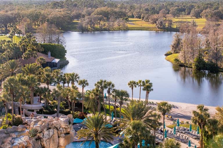 Aerial view of luxury resort with pool, beach, palm trees, and expansive lake surrounded by natural landscape